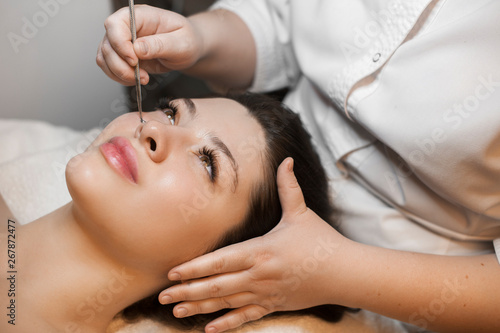 Portrait of beautiful caucasian female with dark hair leaning on a spa bed having face cleaning by a cosmetologist in wellness spa salon.