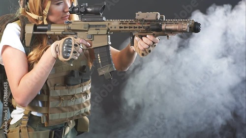 Girl marksman in sniper gear holding sniper rifle in hand aiming at enemy s head in dark smoky background, slow motion photo