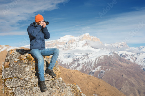 Portrait of a bearded traveler photographer in sunglasses and a cap sits on a rock with mirror camera in his hands against the backdrop of mountains © yanik88