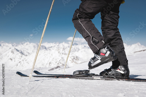 Close-up of the athlete's skier's foot in ski boots rises into the skis against the background of the snow-capped Caucasus mountains on a sunny day. Winter sports concept