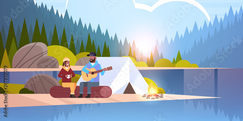 couple tourists hikers relaxing in camp african american man playing guitar for girlfriend sitting on log hiking concept sunrise landscape nature river forest mountains background horizontal