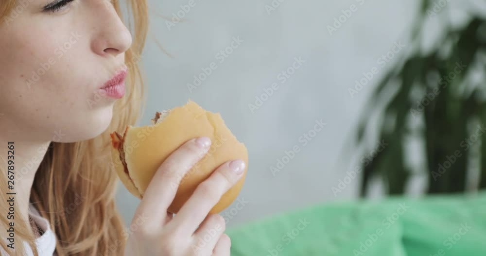 Portrait of young smiling woman is eating burger at home, side view, face closeup.