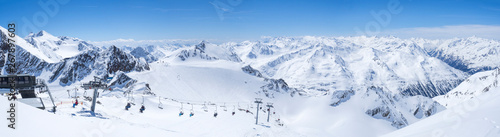 Panoramic landscape view from top of Wildspitz on winter landscape with snow covered mountain slopes and pistes and skiers on chair lift at Stubai Gletscher ski resort at spring sunny day. Blue sky © Kristyna