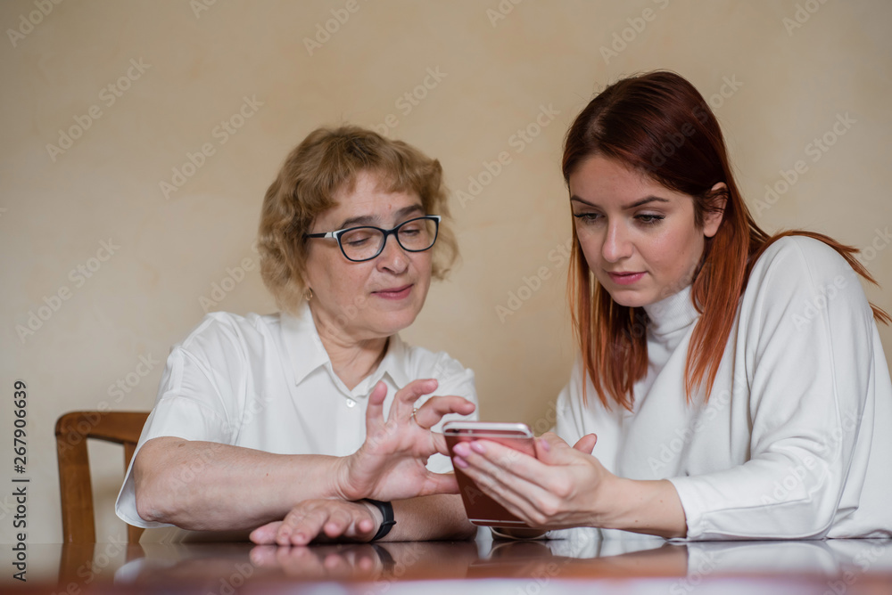 Mother and daughter dressed in light clothes versed in the smartphone. A woman shows how to use a smartphone for her elderly mother Mother and daughter dressed in light clothes versed in the smartphone. A woman shows how to use a smartphone for her elderly mother