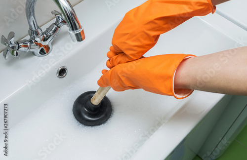 Person in protective orange gloves unblocking a clogged sink with plunger or rubber pump. Close-up, selective focus. 