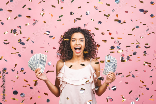Black afro american girl won money. Happy young woman holding dollar currency satisfied isolated over pink background with confetti.