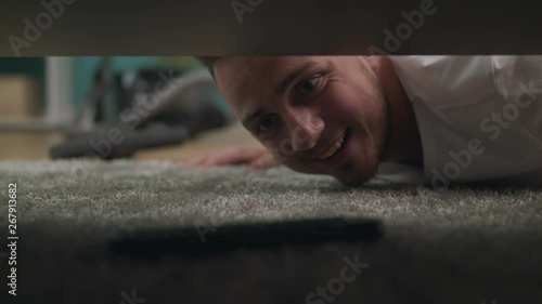 A young guy finds a lost tv remote under the sofa in the living room