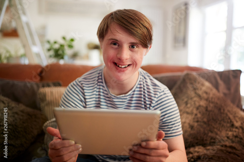 Portrait Of Young Downs Syndrome Man Sitting On Sofa Using Digital Tablet At Home