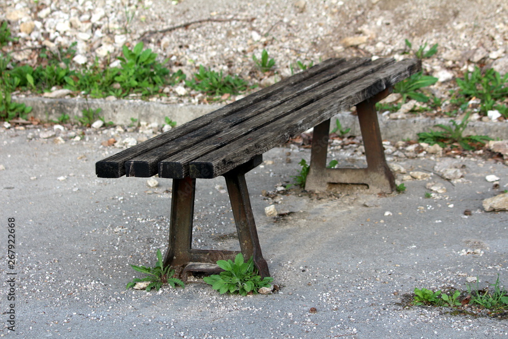 Old wooden public bench with strong rusted metal supports mounted on paved edge of abandoned parking place surrounded with rocks and uncut small plants on warm sunny spring day Old wooden public bench with strong rusted metal supports mounted on paved edge of abandoned parking place surrounded with rocks and uncut small plants on warm sunny spring day
