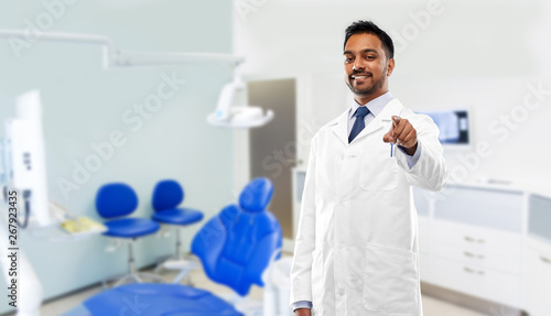 medicine, dentistry and healthcare concept - smiling indian male dentist in white coat pointing to you over dental clinic office background