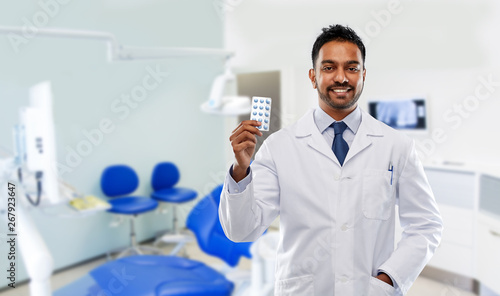 medicine  dentistry and healthcare concept - smiling indian male dentist in white coat with pills over dental clinic office background