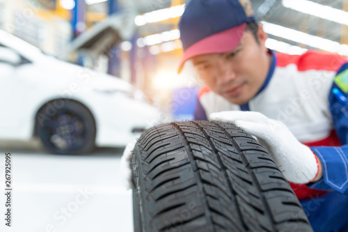 Asian man mechanic in uniform posing on spare wheel, Spare tire car, Seasonal tire change, Car maintenance and service center. Vehicle tire repair and replacement equipment.