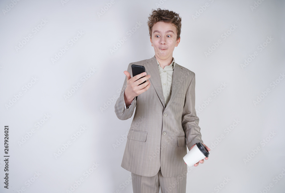 young teenager with surprised emotions in grey business clothes standing with mobile phone and paper cup of coffee on white studio background young teenager with surprised emotions in grey business clothes standing with mobile phone and paper cup of coffee on white studio background
