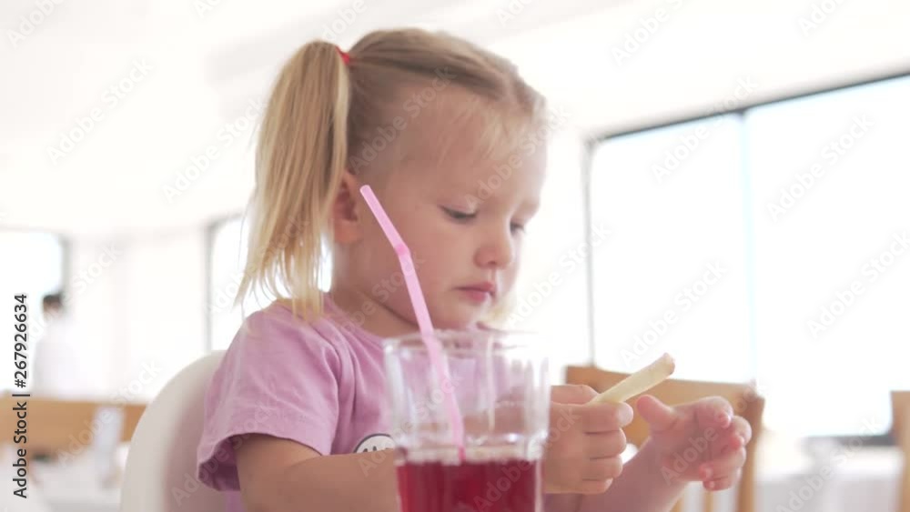 Little girl eating french fries with ketchup in a cafe. The child moistens potatoes in ketchup and chews it deliciously Little girl eating french fries with ketchup in a cafe. The child moistens potatoes in ketchup and chews it deliciously