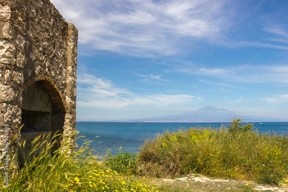 Brucoli view of sea from coast, green grass near an old building, blue sky and the distant Etna