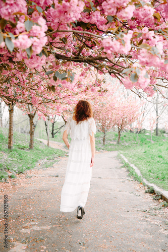A young woman in a white dress in the spring park with a pink sakura tree in blossom