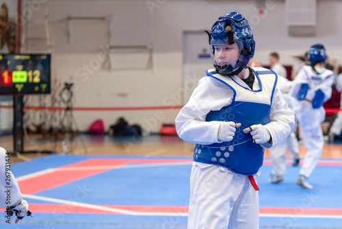 a girl participating in Taekwondo competitions in a blue protective vest and a protective helmet is a duel in Taekwondo against the background of Taekwondo competitions close-up