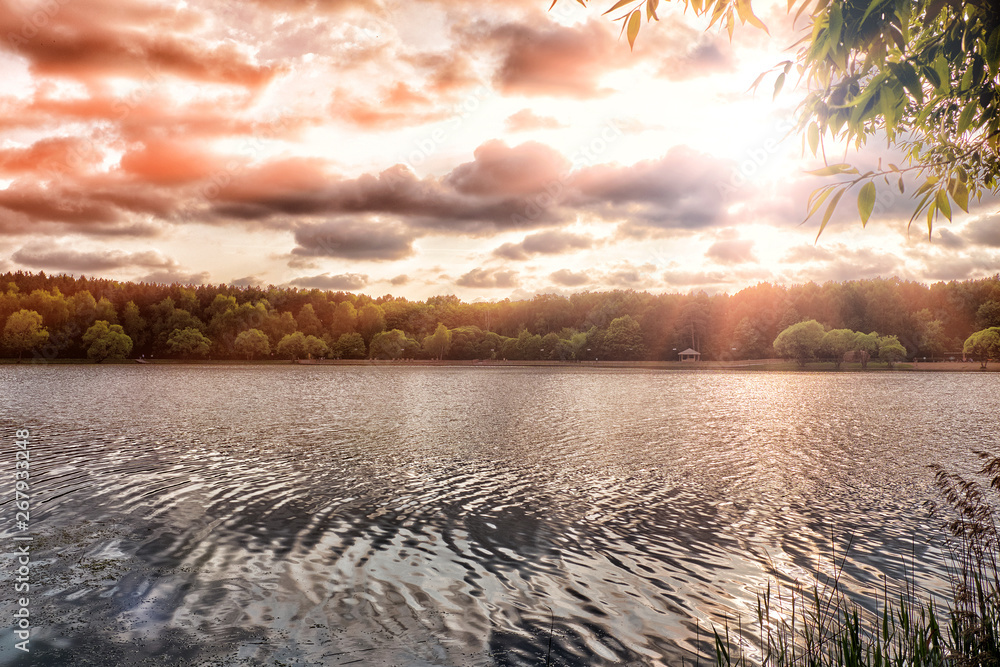 sunset at city park with pond on summer evening ground landscape view of outdoor scenery reflection in lake water with sun rays light shines through red dramatic clouds sky nature environment concept