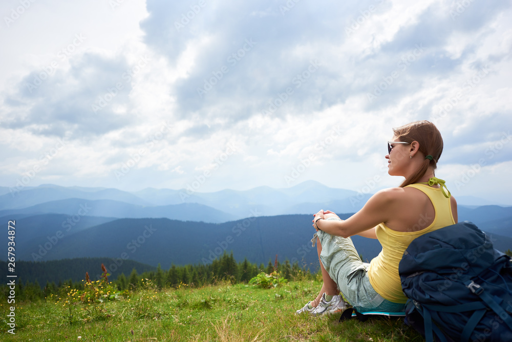 Back view of happy woman backpacker sitting and resting on grassy hill with backpack, wearing sunglasses. Female traveler enjoying summer cloudy day in the mountains. Outdoor activity, tourism concept