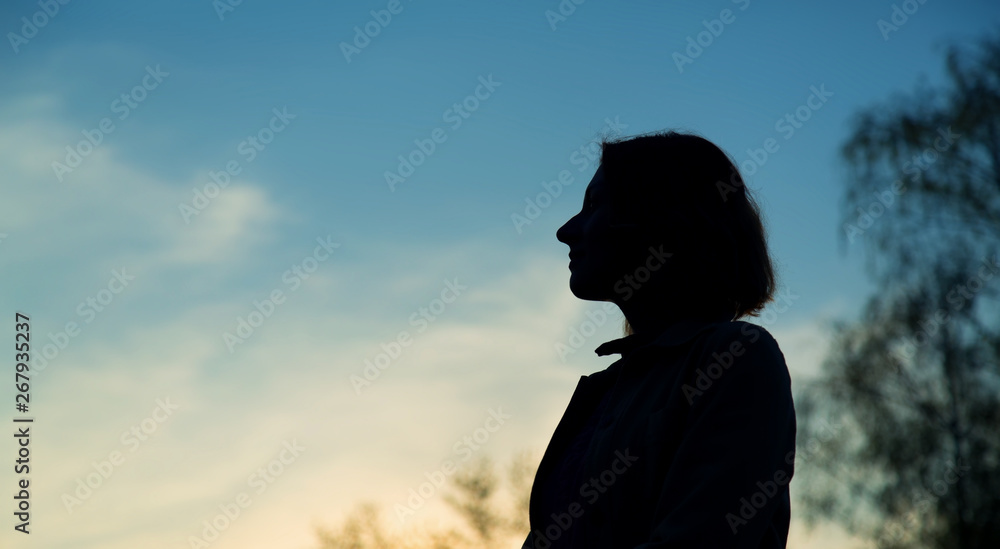 Silhouette of a woman against the backdrop of the sunset, the forest and the blue sky.