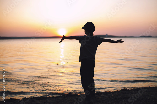 boy at sunset stands on on the beach and raised his hands up