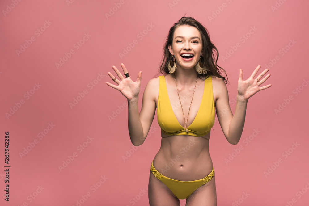 Naklejka premium front view of excited girl in yellow swimsuit laughing and gesturing isolated on pink