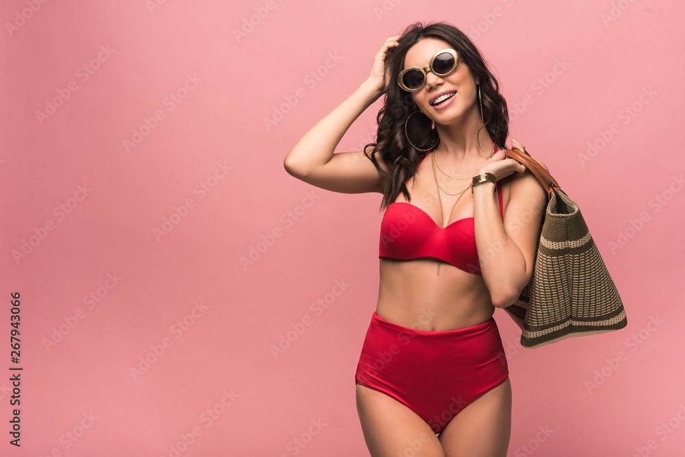 Obraz premium smiling attractive woman in swimsuit and sunglasses holding beach bag isolated on pink
