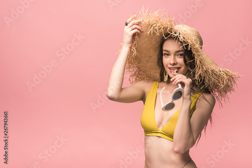 smiling girl in yellow swimsuit and straw hat holding sunglasses isolated on pink