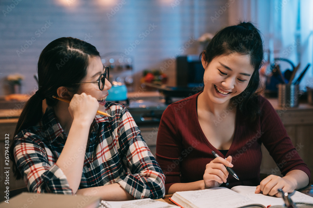 Laughing two female student learning with friend indoors at late night in dormitory kitchen. young college girls smiling having fun chatting while doing homework in midnight. women stay up late.