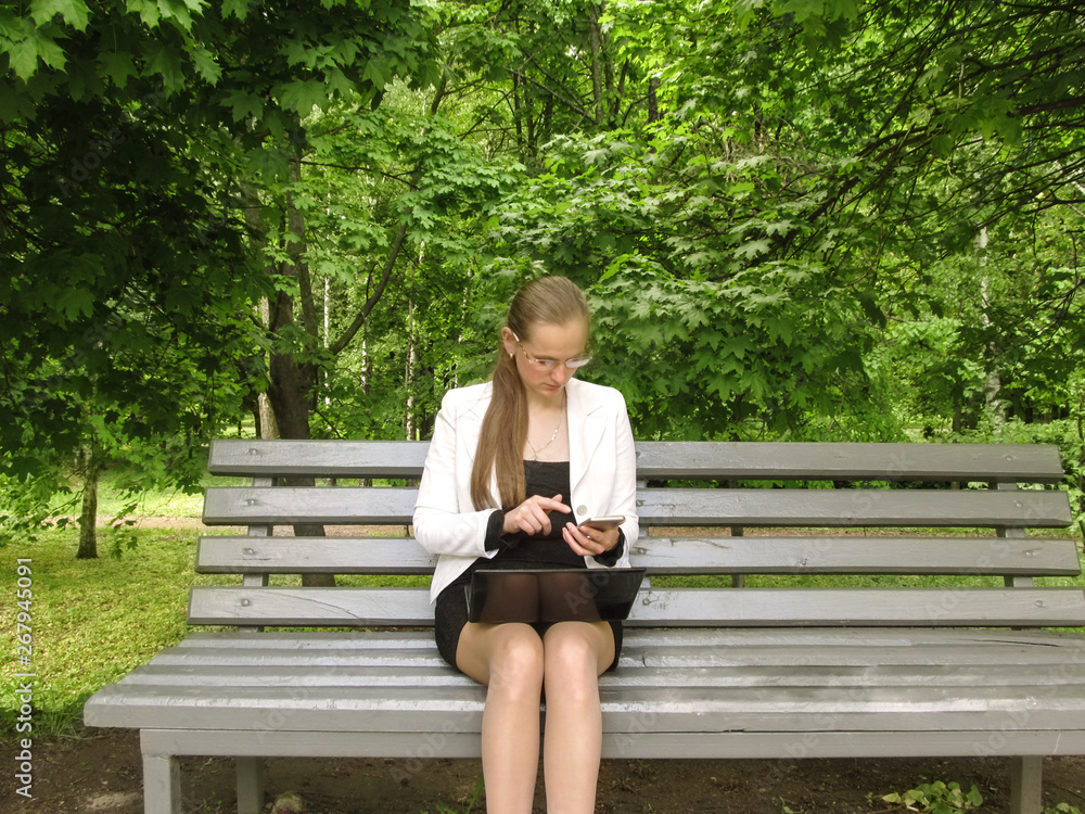 Obraz premium The girl in glasses dials the number on the screen of the smartphone sitting on a bench in a park. Working with technology outdoors concept, businesswoman with mobile and laptop