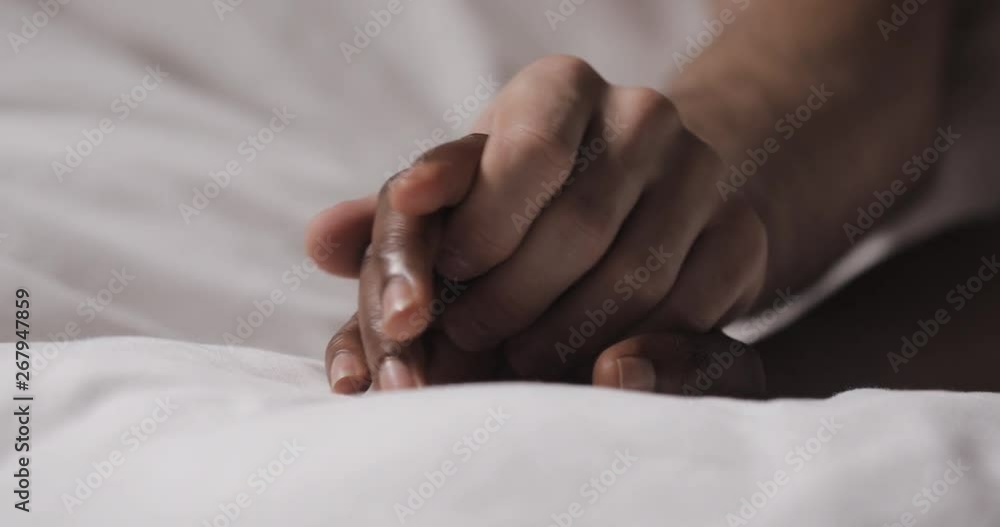 Multi-ethnic couple in love lying in bed together. Close up. Womans hand is lying on a bed, mans hand is caressing it and grabbing. Multi-ethnic couple in love lying in bed together. Close up. Womans hand is lying on a bed, mans hand is caressing it and grabbing.