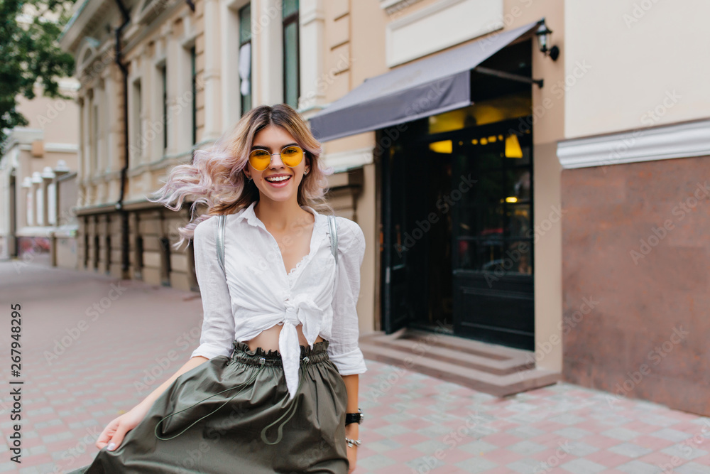 Attractive curly woman with sincere smile playing with her long skirt while walking down the street. Adorable blonde girl with hair waving laughing and posing outdoor near restaurant.