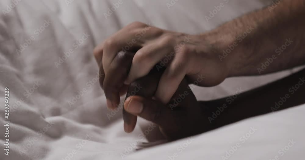 Multi-ethnic couple in love lying in bed together. Close up. Womans hand is lying on a bed, mans hand is caressing it and grabbing. Multi-ethnic couple in love lying in bed together. Close up. Womans hand is lying on a bed, mans hand is caressing it and grabbing.