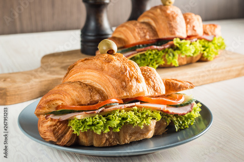 Photo of tasty and fresh croissant sandwich with salad, ham, cheese, tomatoes on wooden background. Morning breakfast concept. Healthy and fast food.