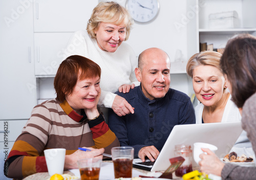 Smiling aged friends are sitting at table with notebook and talking about project