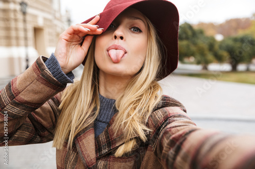 Cheerful blonde woman wearing autumn coat walking outdoors, taking a selfie