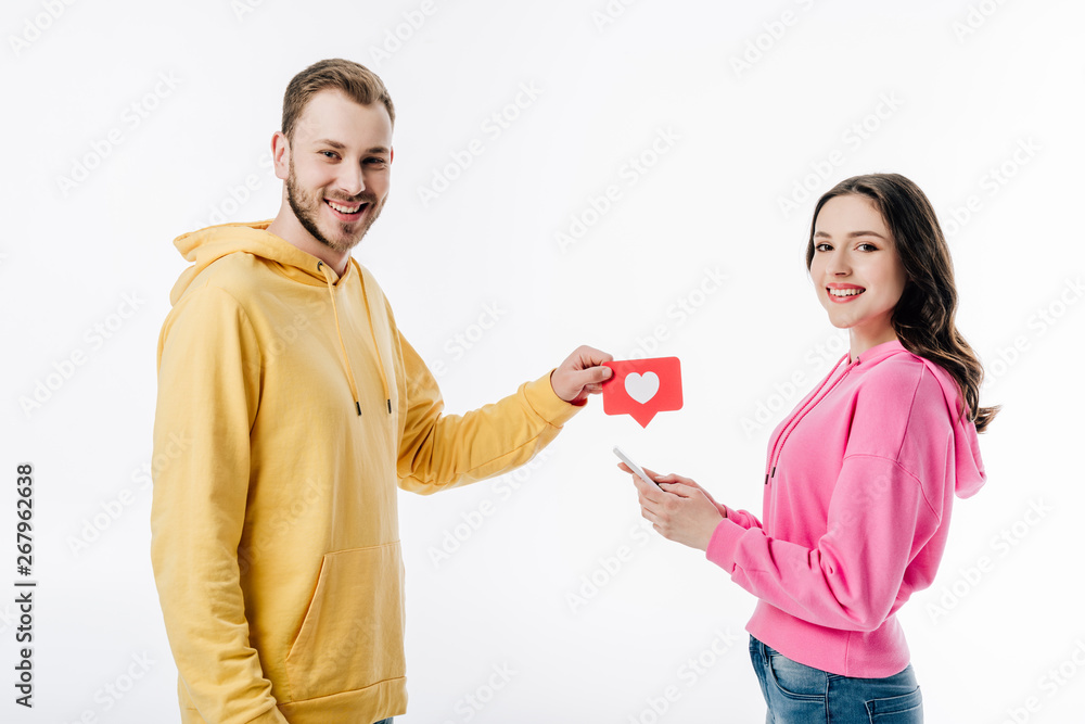 smiling handsome man giving red paper cut card with heart symbol to pretty girl using smartphone isolated on white smiling handsome man giving red paper cut card with heart symbol to pretty girl using smartphone isolated on white