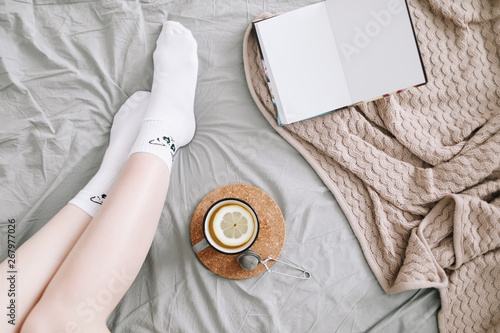 Cozy flatlay of woman legs in bed with a book and a cup of lemon tea. Cozy morning at home. 
