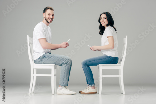 smiling man and woman in white t-shirts and blue jeans sitting on chairs and using smartphones on grey background