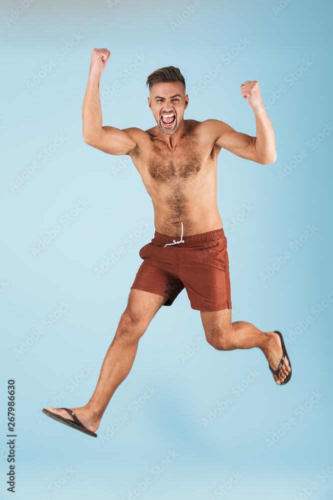 Excited happy adult man in swimwear posing isolated over blue wall background jumping make winner gesture.