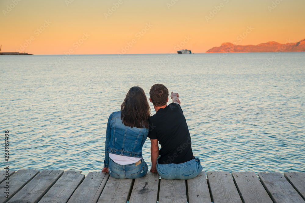 Couple in love sitting together on the pier by the sea. Colorful sunset landscape around