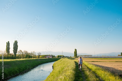 young woman riding bike in italian countryside along "sentiero degli ezzelini" path in Veneto region with river and mountains during summer