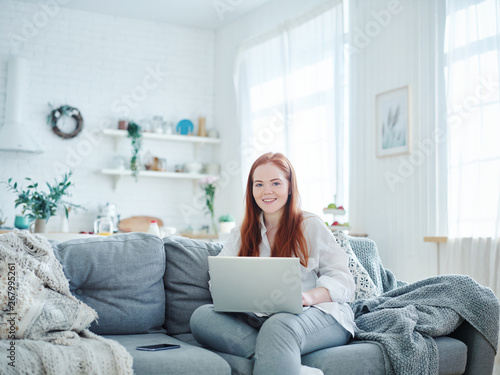 Portrait of beautiful red-haired woman looking at camera and smiling while working on laptop on cosy sofa at home, copy space on top