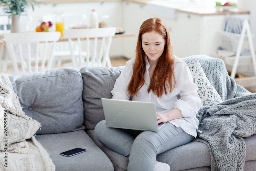 Portrait of beautiful red-haired businesswoman working on laptop sitting on cosy sofa at home, smartphone placed nearby Portrait of beautiful red-haired businesswoman working on laptop sitting on cosy sofa at home, smartphone placed nearby