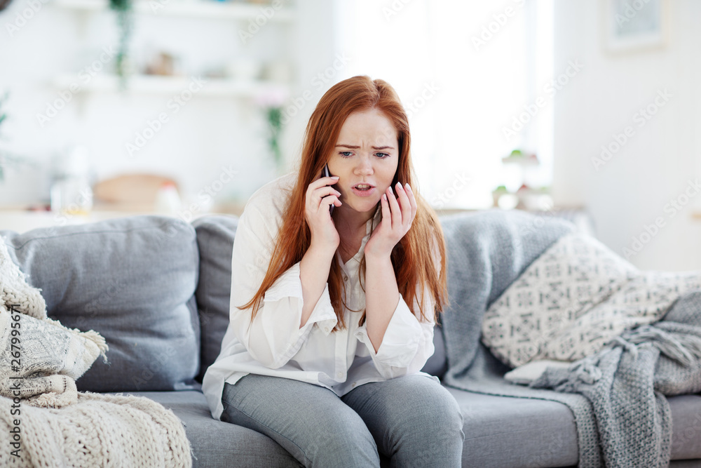 Worried young woman sitting on sofa at home and having negative phone talk, hearing bad news or arguing Worried young woman sitting on sofa at home and having negative phone talk, hearing bad news or arguing