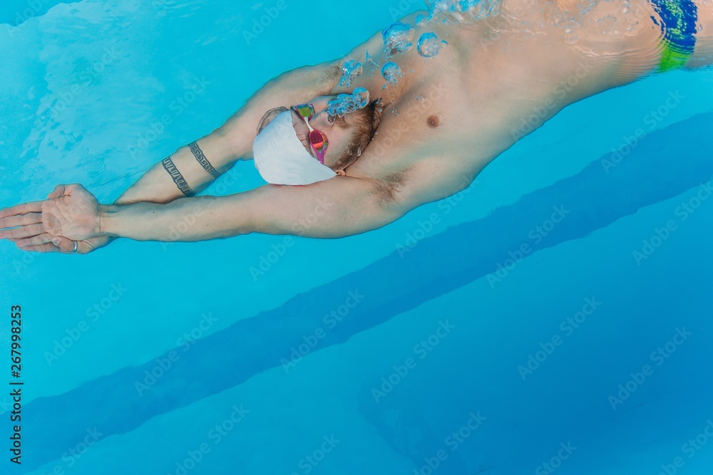 Swimmer in underwater goggles and swimming cap swims underwater in the outdoor pool on a summer day close-up