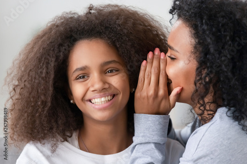 African American mother whispering to smiling teen daughter ear
