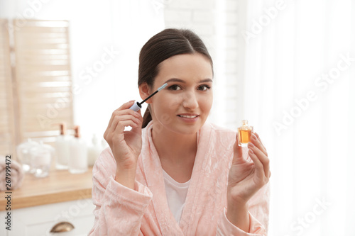 Beautiful woman applying oil onto her eyelashes indoors