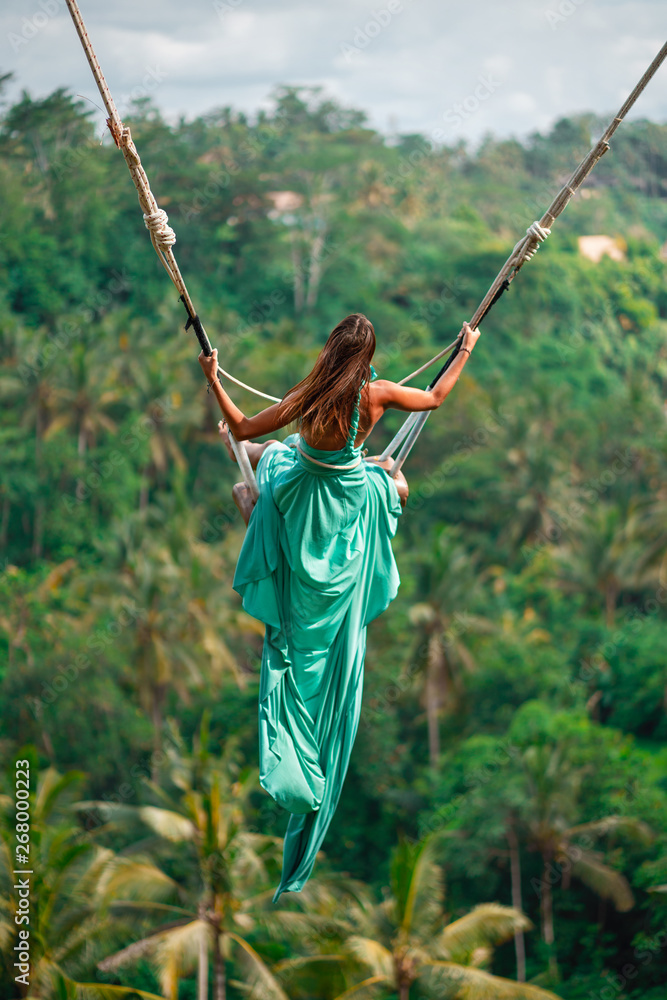 Tanned young woman riding on a long swing in long turquoise dress. Island  of Bali. Tropical forest on the background. Travel and joy Photos | Adobe  Stock