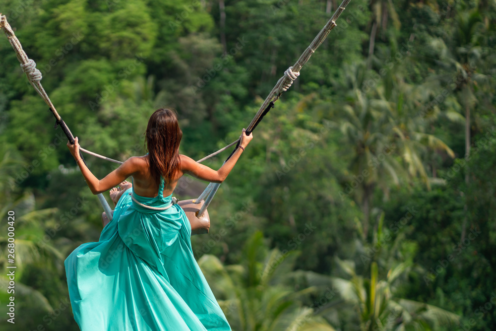 Woman in long turquoise dress swinging in the jungle, Bali. Copy space
